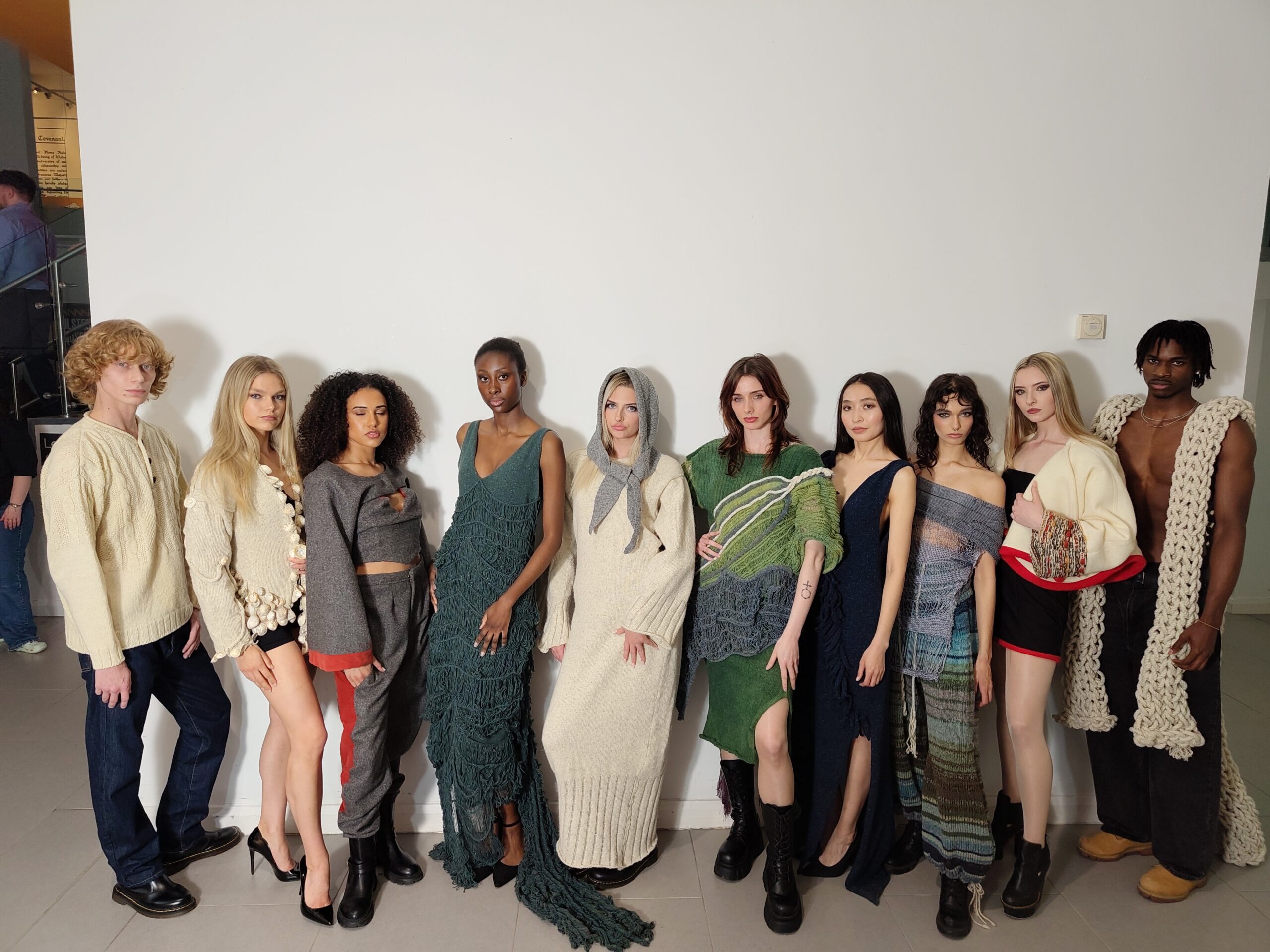 Here's alt text for this image: Alt text: "Ten models of diverse ethnicities stand in a line against a white wall, wearing handcrafted knitwear and textile garments in a palette of cream, grey, teal, green, and multicolour. Pieces include heavily textured fringe dresses, chunky knit sweaters, draped scarves, and woven skirts